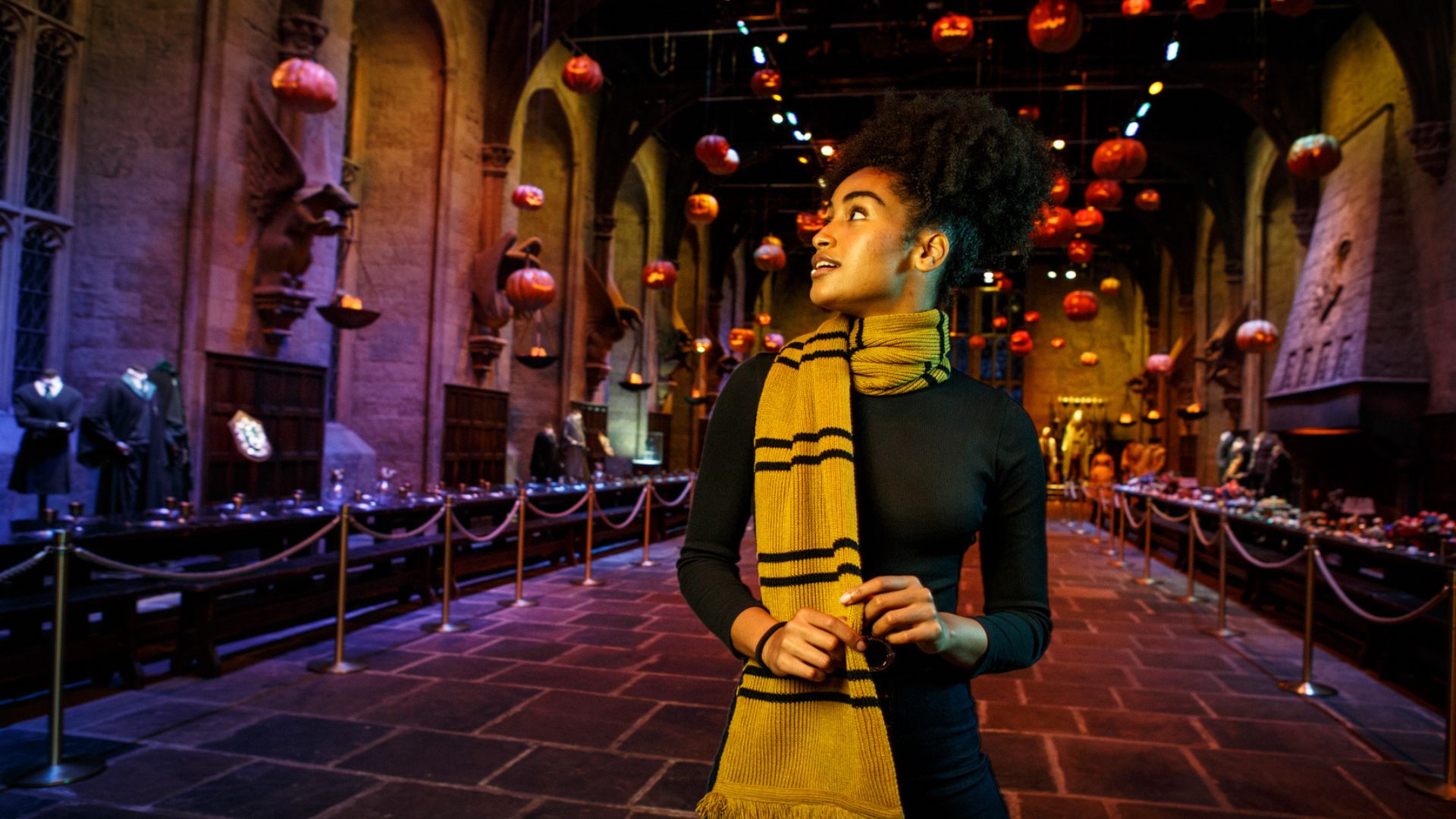 It's Halloween at Hogwarts and you're invited to explore the spooky Dark Arts exhibit. Image courtesy of Golden Tours. A person stands in the Great Hall of Hogwarts, decorated with floating jack-o'-lanterns for Halloween. The hall features long tables set for a feast, stone walls with gothic architecture, and statues of winged creatures. The person is wearing a yellow and black striped scarf and looking to the side, capturing the magical ambiance