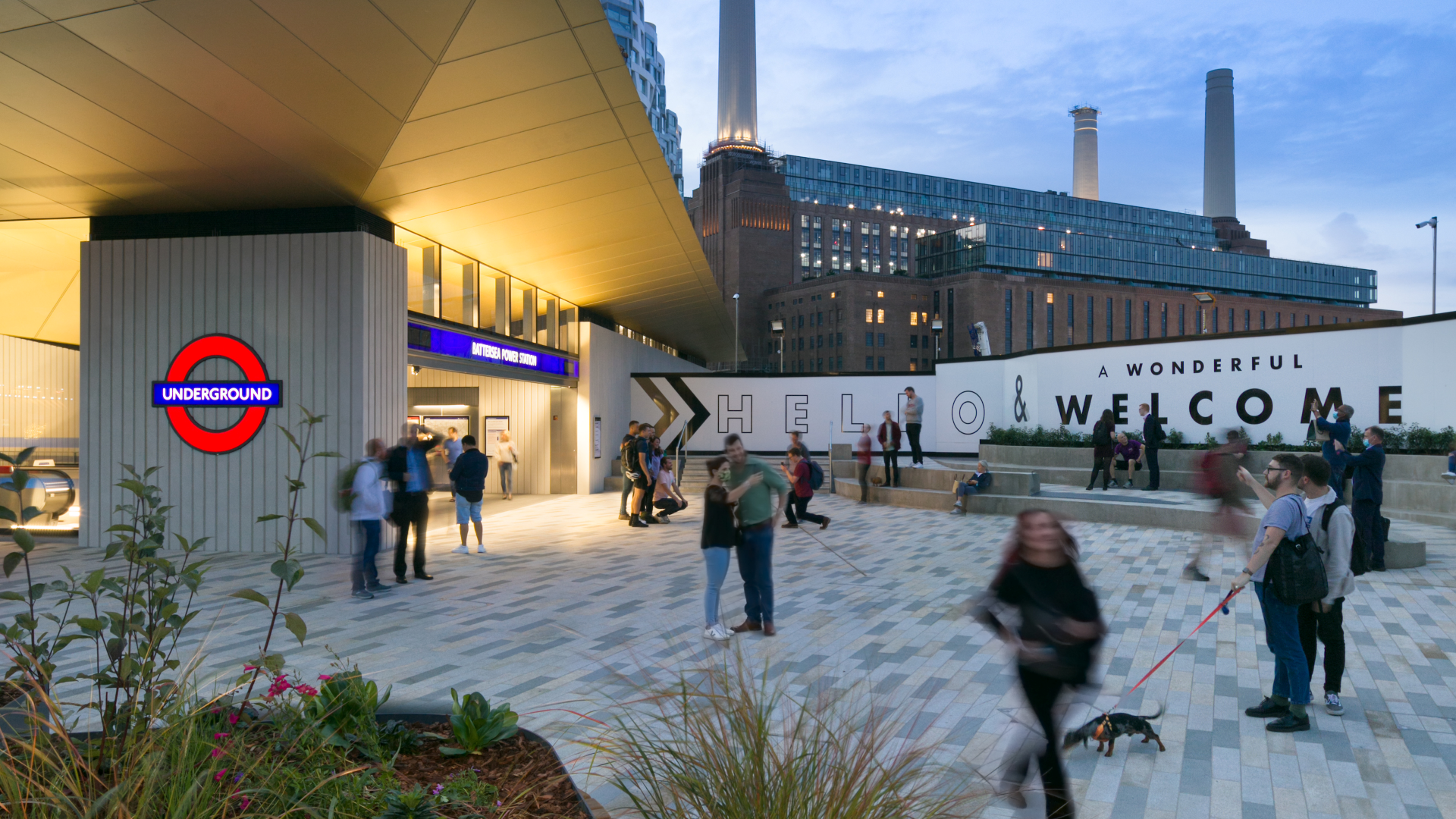 Discover a bustling food, shopping and entertainment hub at the iconic Battersea Power Station. Credit: Brendan Bell. Image courtesy of Brendan Bell. Battersea Power Station Tube station in the evening with people walking around in the foreground and the iconic Battersea Power Station with lights on in the background