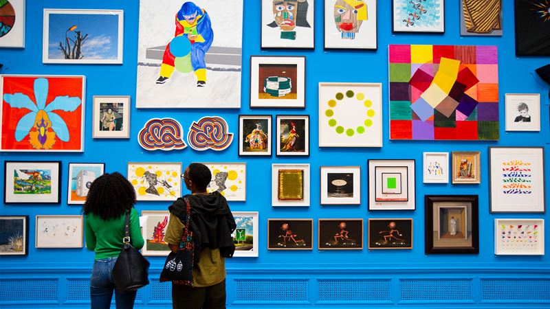 Installation view of the Summer Exhibition 2024 at the Royal Academy of Arts in London, 18 June - 18 August 2024. Image courtesy of Royal Academy of Arts, London / David Parry. A couple looking at a wall covered in different artworks at the Summer Exhibition
