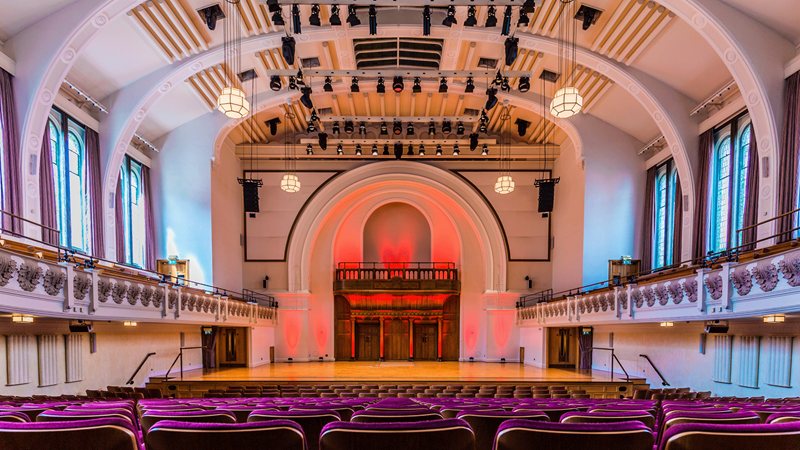 Listen to musical performances at Cadogan Hall. Credit: TODD Creative Services. Image courtesy of TODD Creative Services. View from the seats facing the stage at Cadogan Hall with red lights illuminating the stage