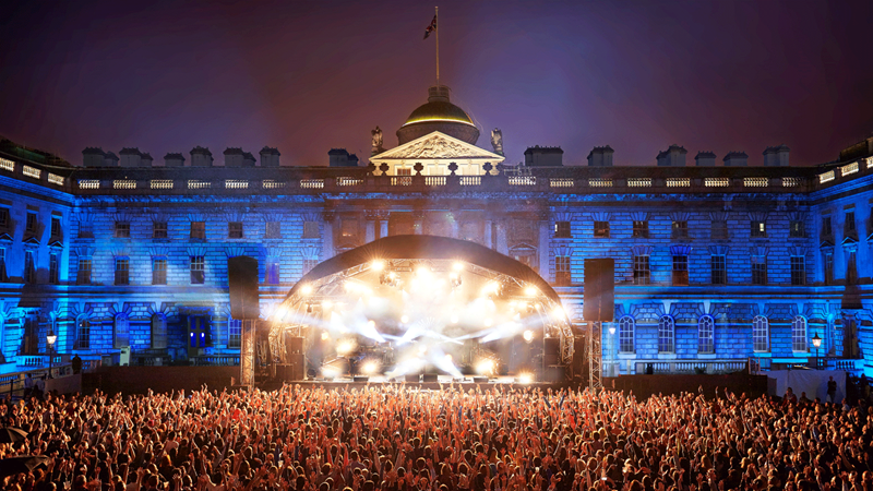 See world-class talent with an iconic backdrop at Somerset House Summer Series. Credit: James Bryant. Image courtesy of James Bryant. White lights illuminate the stage and blue lights light up Somerset House behind it with the crowd in the foreground