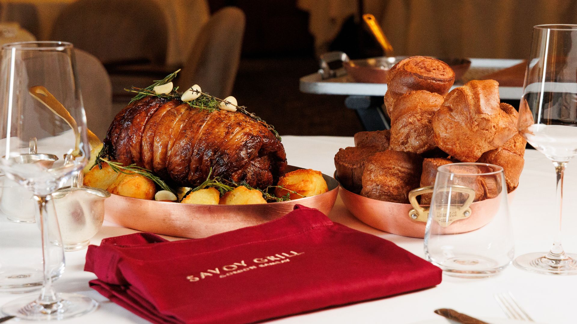Take centre-stage with a Host & Roast experience at the Savoy Grill. Image courtesy of Gordon Ramsay Restaurants. Saddle of lamb surrounded with roast potatoes on a dish with pile of Yorkshire puddings next to it and Savoy Grill red cloth in front.