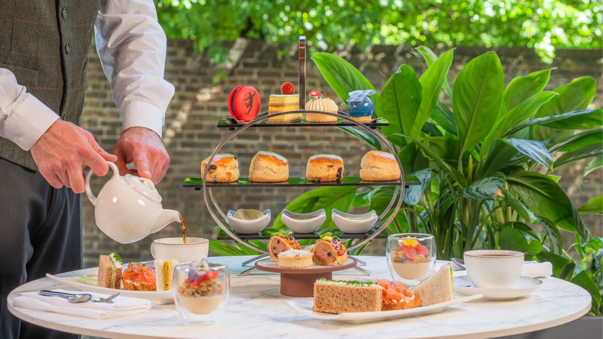 Tuck into The Beautiful Game Afternoon Tea at Origin Kensington. Image courtesy of the Royal Garden Hotel. The Beautiful Game Afternoon Tea with football theme on table with waiter pouring from a teapot.
