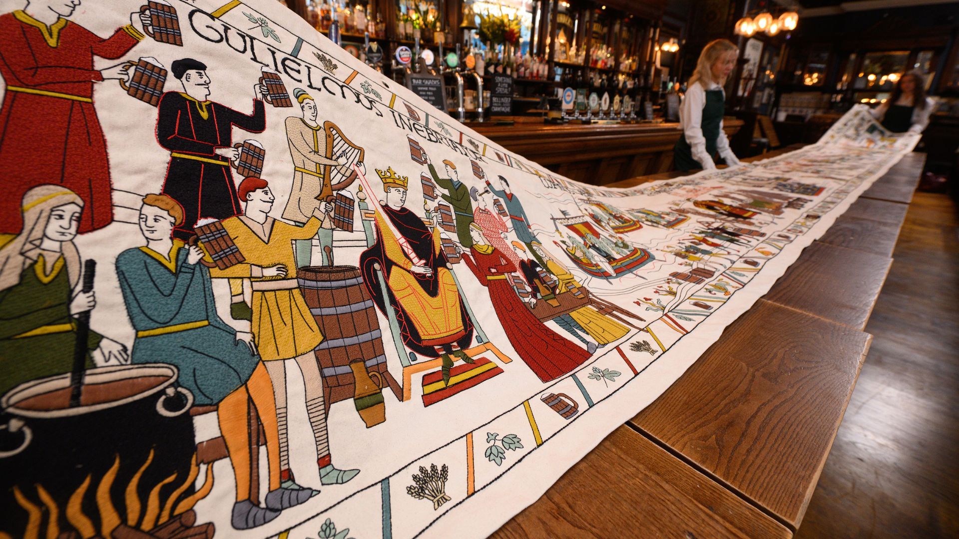 See the Beer Tapestry exhibition this weekend, a playful twist on the historic Bayeux Tapestry at the British Museum. Image courtesy of Pink Neon Creative. a long wooden table in a pub, with a long, embroidered tapestry detailing the history of beer, held by people in suits and white gloves