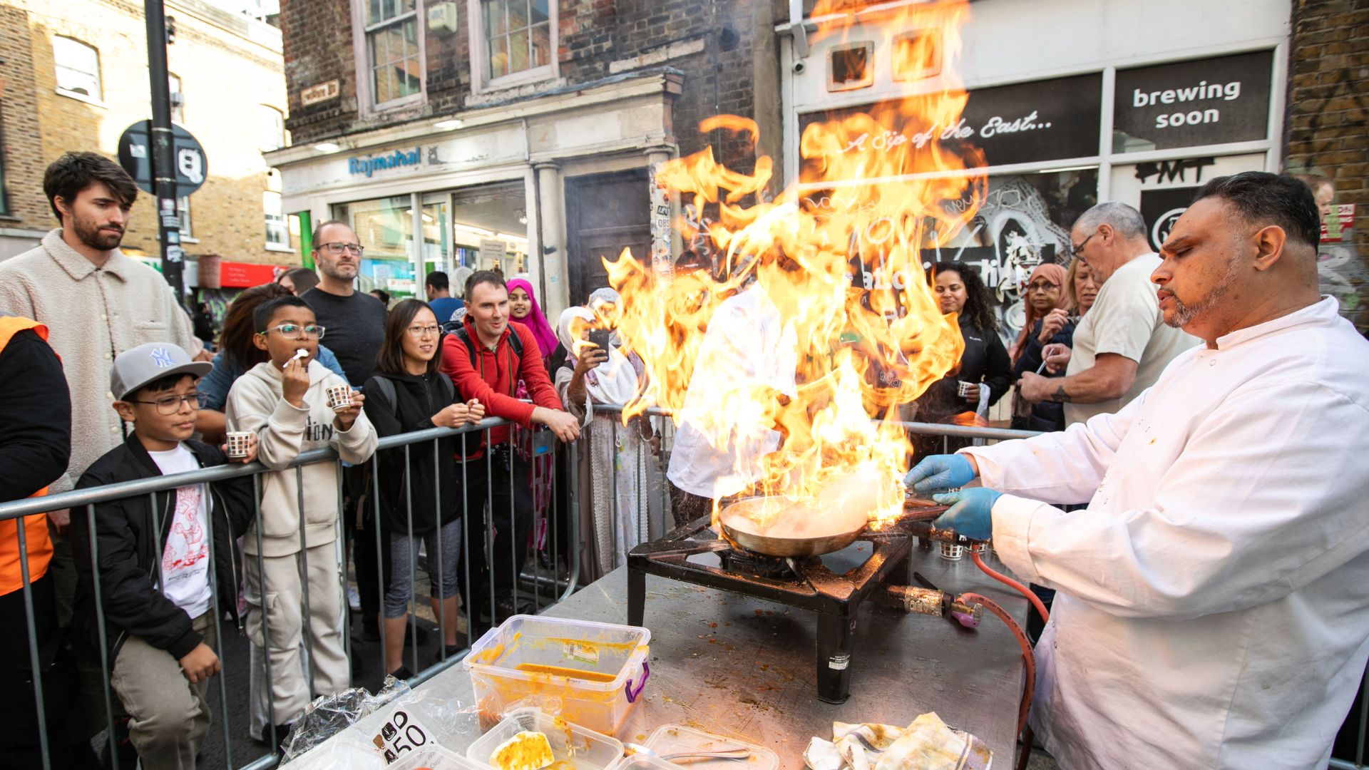 Celebrate Brick Lane's food, culture and community at the Brick Lane Curry Festival. Image courtesy of Brick Lane Curry Festival. a chef set up on a cooking table outside on a street, holding a pan with flames, whilst of smile and watch from behind a barrier