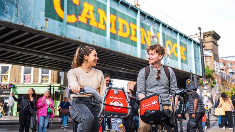 Tour London's famous sights and landmarks on a Santander Cycles bike. Credit: Golden Tours. Image courtesy of Golden Tours. A man and women smiling at each other while sat stationary on Santander Cycles bikes with the famous Camden Lock Bridge in the background