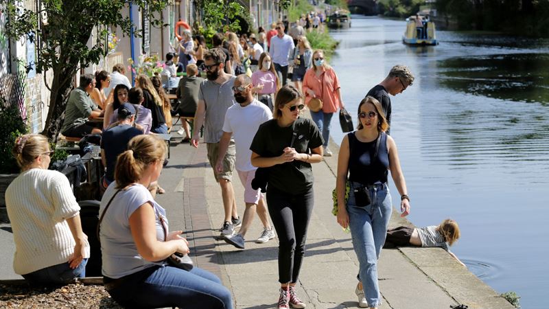 Stroll Regent's Canal during the summertime for picturesque views. Credit: Shutterstock. Image courtesy of Shutterstock. A photo of lots of people dressed for summer walking and sat along Regent's Canal as the sun shines down on them