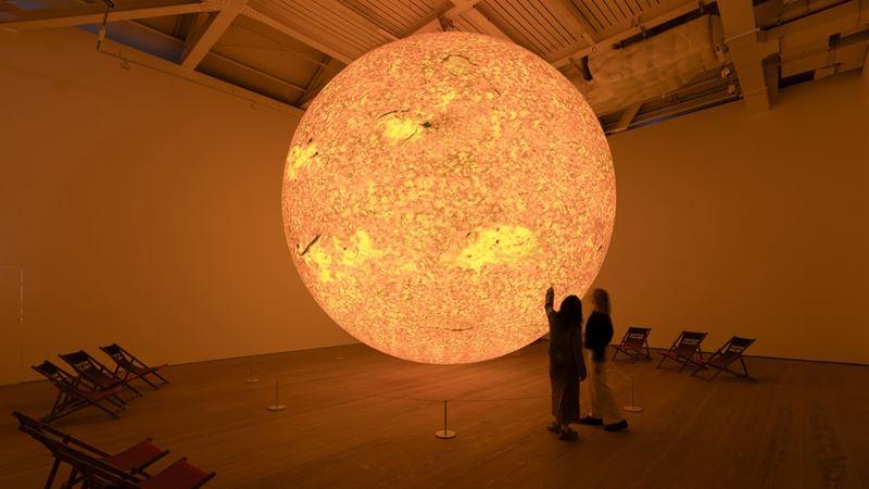 Discover Helios by Luke Jerram at Saatchi Gallery's The Sun and The Moon exhibition. Photo credit: David Owens Photography. Image courtesy of Saatchi Gallery. Large-scale sculpture of the sun with two people standing next to it in gallery room.