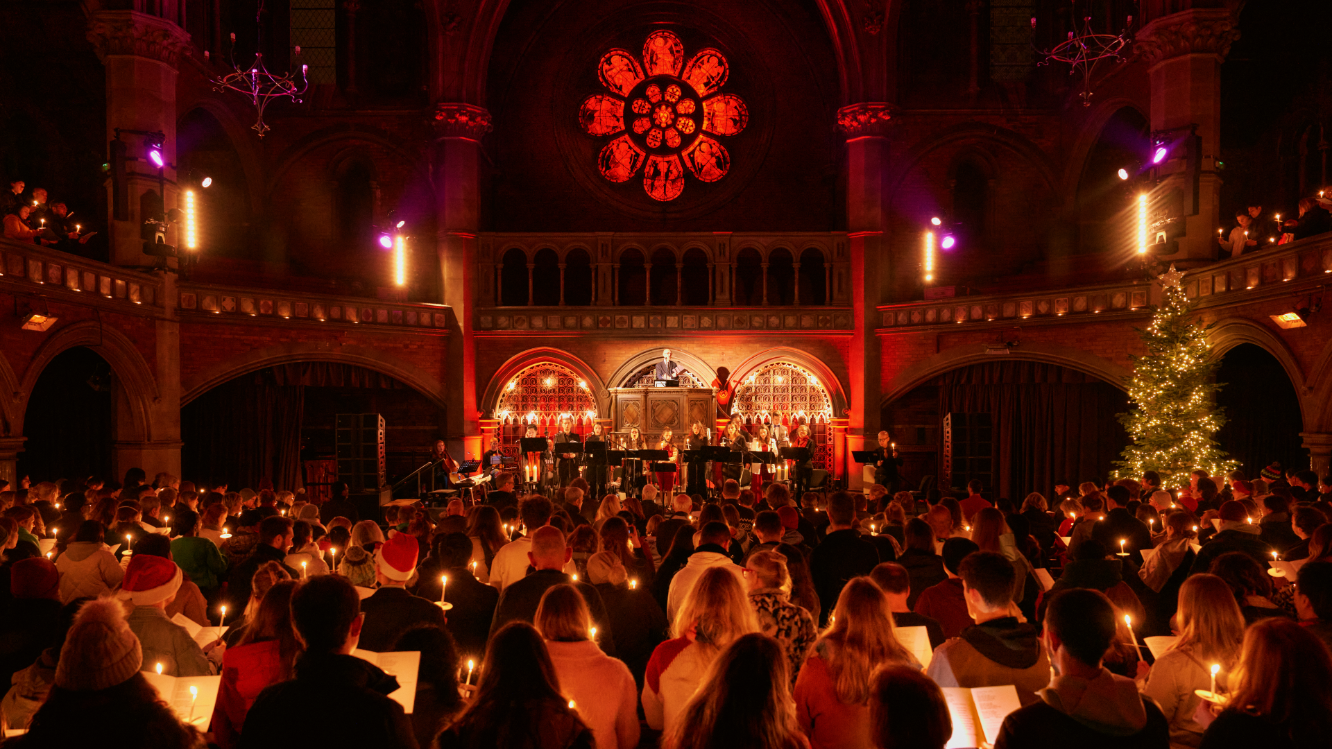 Christmas carols at Union Chapel in Islington. Image courtesy of Union Chapel/Emile Holba. Crowds of people stand on across two levels at Union Chapel as a round red stained glass window illuminates the whole room with performers and musicians stood on the stage