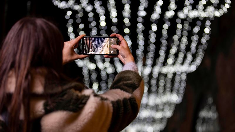 Snap photos of the twinkling ceiling of light above Camden Market during Christmas. Credit: David Parry. Image courtesy of Camden Market. A photo of a woman taking a photo on her phone of a canopy of twinkling lights above Camden Market