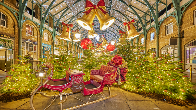 Don't miss some of London's top Christmas decorations at Covent Garden. Credit: Covent Garden. Image courtesy of Covent Garden. A red sleigh with gold details in the Covent Garden market surrounded by Christmas trees with twinkling lights with giant gold bells with bows hanging above