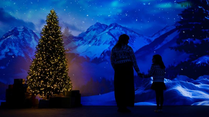 Immerse yourself in a wintry wonderland during the festive season at Frameless. Credit: David Parry. Image courtesy of Frameless. A photo inside Frameless gallery with a Christmas tree and snowy mountains on screen along with an outline of a mother and young child