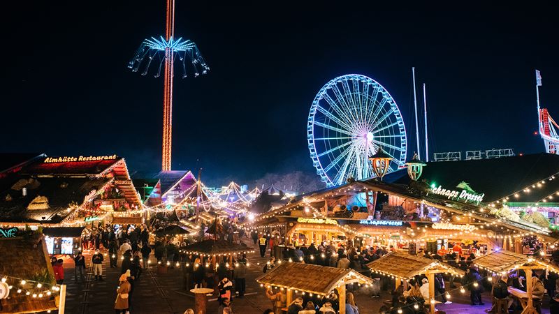 Winter Wonderland Christmas market stalls. Credit: Winter Wonderland. Image courtesy of Winter Wonderland. Twinkling string lights hang above people perusing wooden Christmas market stalls.