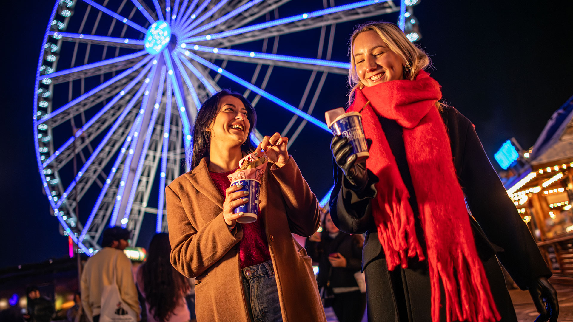 Visit Winter Wonderland during the festive season, with one of the capital's biggest Christmas markets. Image courtesy of London & Partners/Michael Barrow. Two young women drinking hot chocolate in Winter Wonderland, with the Great Wheel in the background.