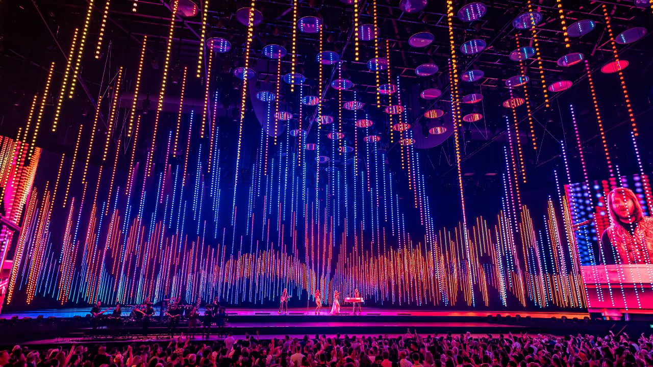 ABBA Voyage performance of Dancing Queen at London's ABBA Arena. Image courtesy of Johan Persson / ABBA Voyage. ABBA Arena stage lit up in pink, orange and purple lights during a live performance of ABBA Voyage in London.
