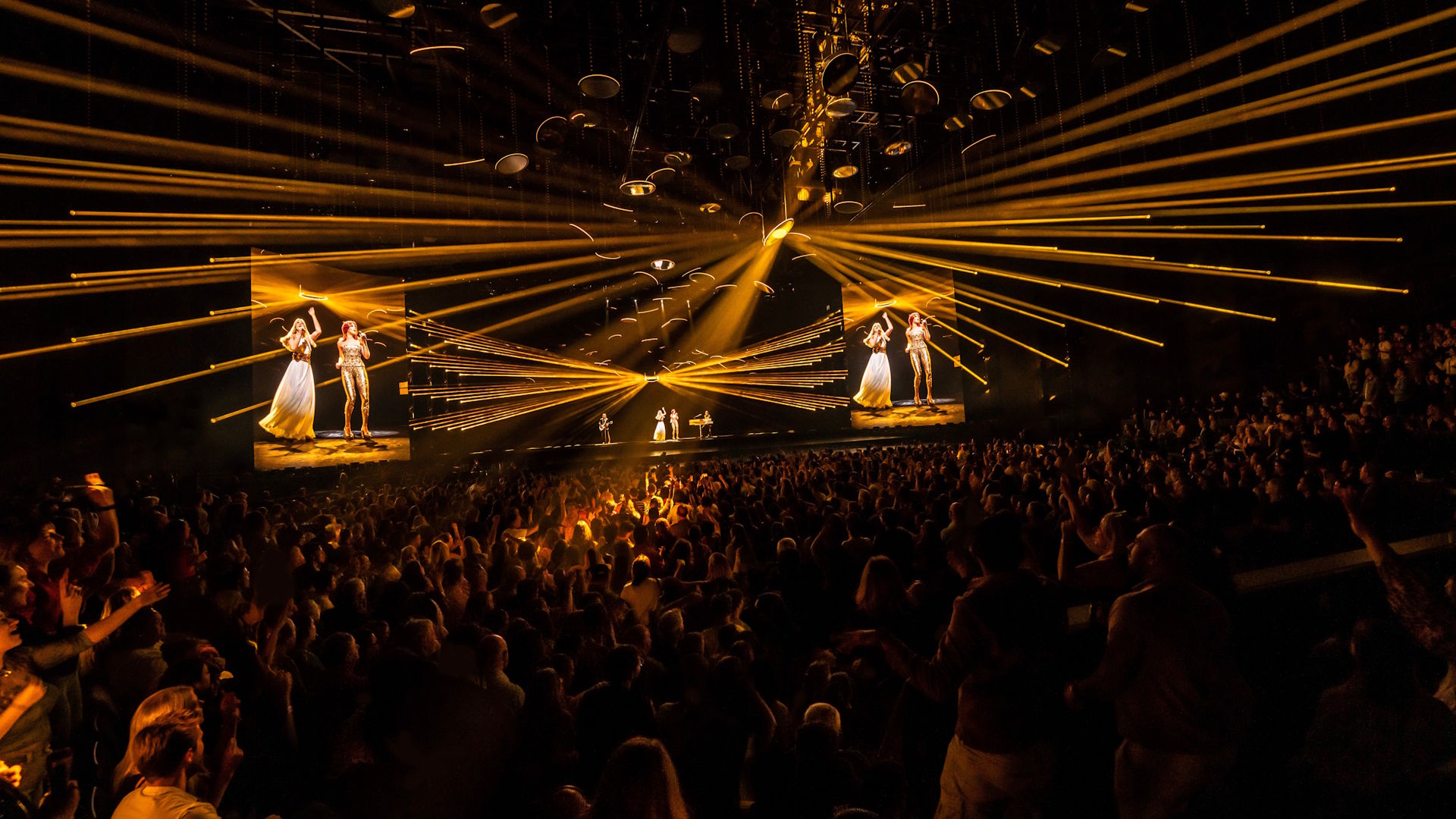 See the performance of hits such as Money, Money, Money at ABBA Voyage, a concert like no other. Image courtesy of ABBA Voyage / Johan Persson. A crowd looks on in the dark as numerous yellow lasers shine out from the stage, with digital avatars of ABBA band members performing on stage.