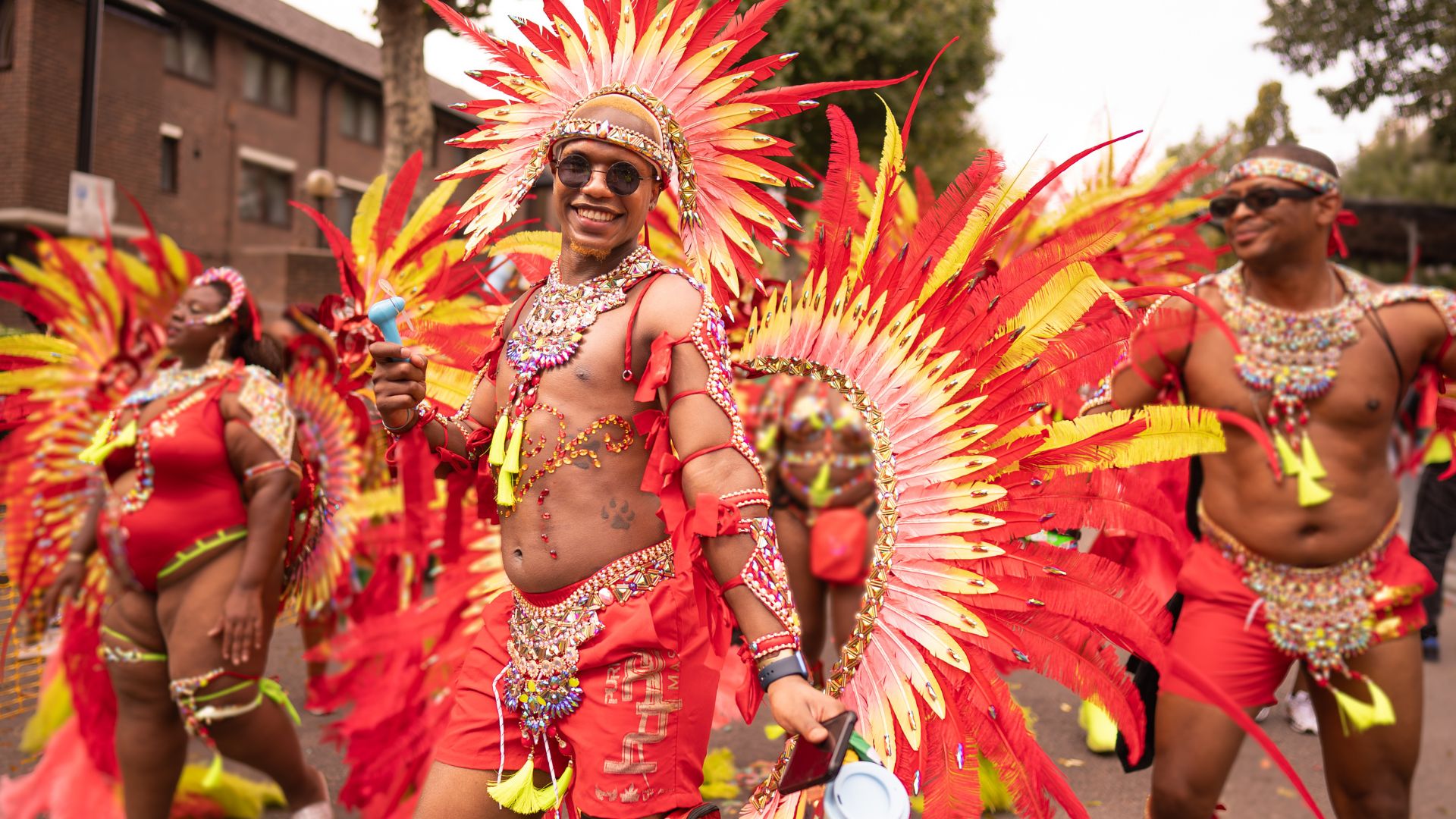 Experience the vibrant atmosphere of Notting Hill Carnival as masquerade performers fill the streets with colour and creativity. Image credit: London & Partners/Peter Cohen. A carnival performer smiles while wearing an elaborate red, orange and gold feathered costume during Notting Hill Carnival. Image credit: London & Partners/Peter Cohen.