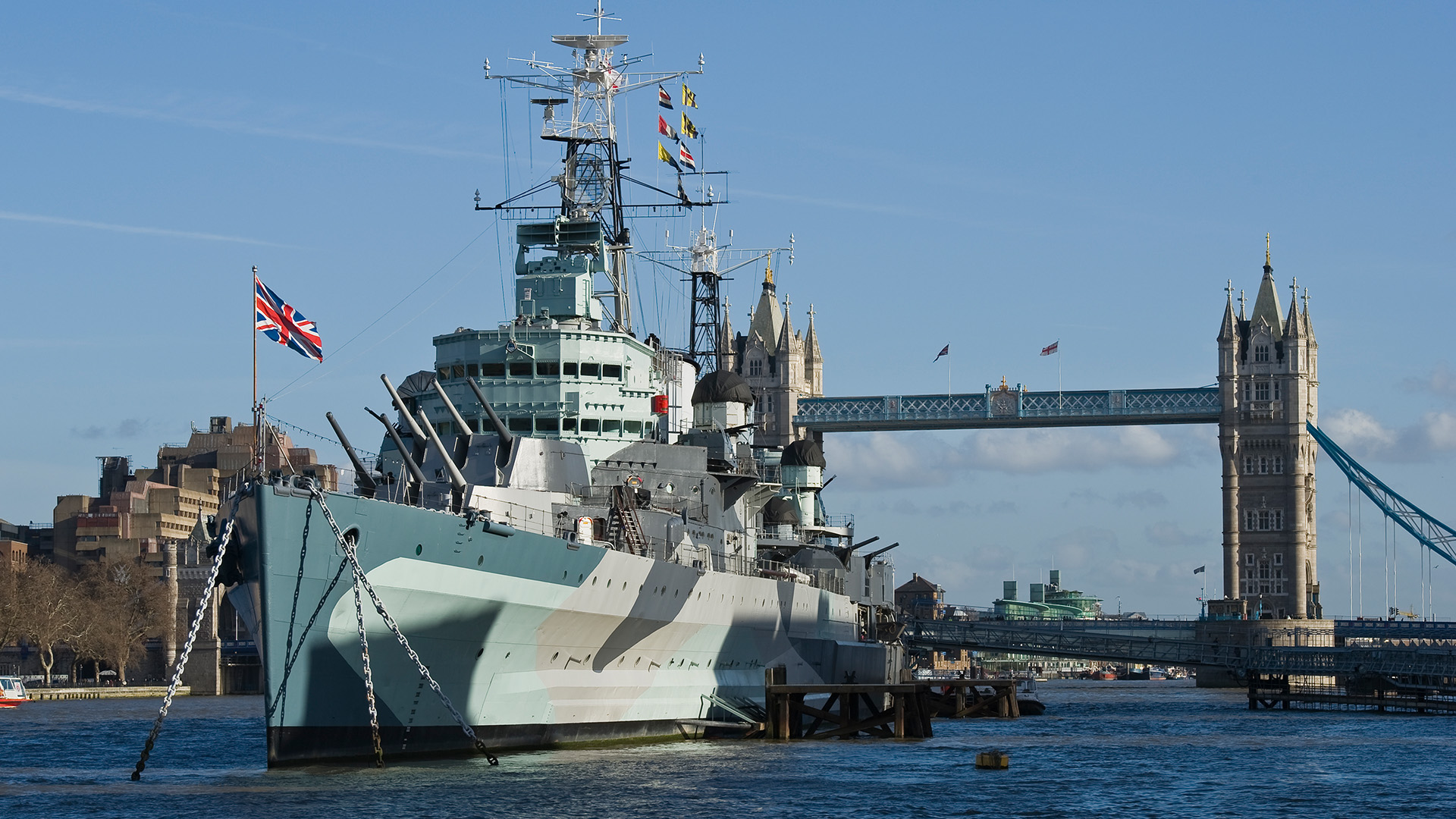 Take a tour of the famed HMS Belfast on the Thames. Image courtesy of IWM/Richard Ash.. A battleship sits atop the water with a bridge and blue sky in the background.