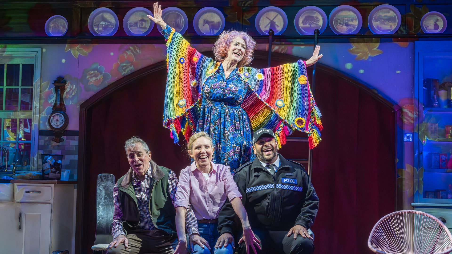 See the heartwarming comedy Allegra, starring Maureen Lipman, bursting with joy and featuring unforgettable sing-along moments. Photo credit: Marc Brenner. Wearing a blue dress and a rainbow-coloured knitted poncho, Maureen Lipman stands on a table, raising her hands in the air. The other cast members are seated and smiling at the audience.