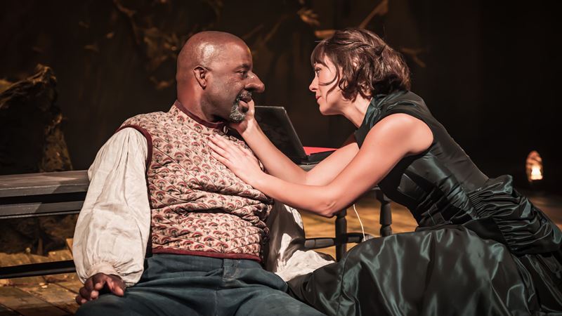 See Adrian Lester star in a West End remake of Edmund Rostand’s Cyrano de Bergerac. Image courtesy of London Theatre Direct. Adrian Lester and Susannah Fielding gaze at each other while seated during a performance of Cyrano de Bergerac.