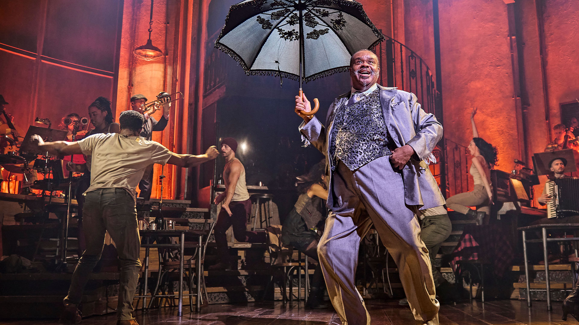 See Hadestown, the Tony Award-winning musical that blends folk, pop and New Orleans jazz, set to the intertwining love stories of the underworld. Image courtesy of London Theatre Direct/ ©-Brinkhoff-Moegenburg. Clive Rowe, who plays Hermes in Hadestown, stands on stage wearing a silver three-piece suit and holding a black umbrella. Cast members can be seen dancing in the background.