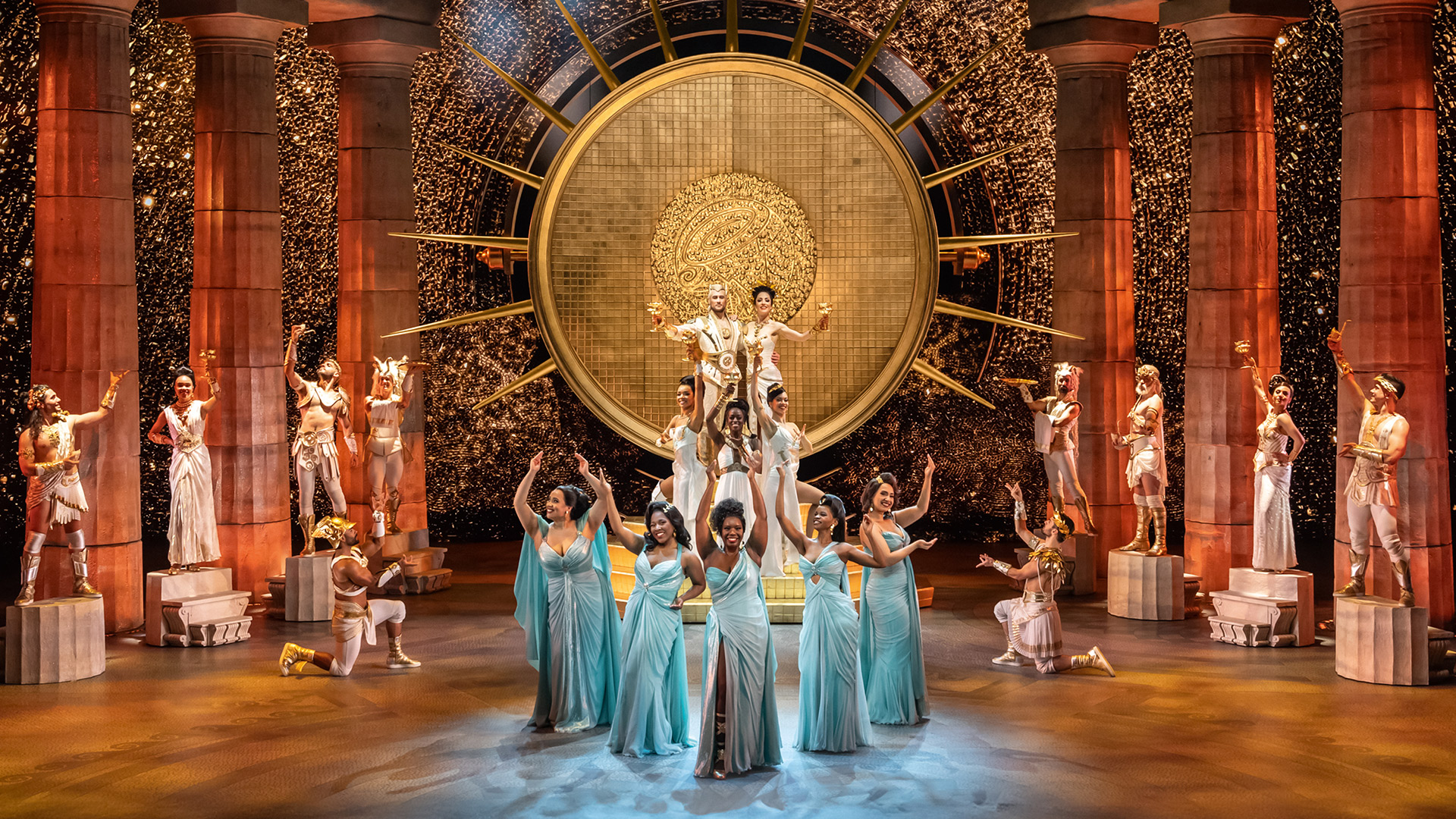 See Disney's highly anticipated musical production of Hercules, which will premiere in the West End in 2025. Image courtesy of Original Hamburg Company ©Disney, Photo by Johan Persson Women in long light blue dresses are at the centre of the stage, with other members of the production in white in the background.