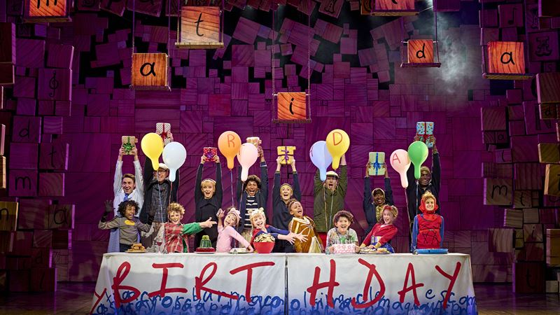 See the RSC's award-winning production of Matilda The Musical, a stage adaptation of Roald Dahl's bestselling book. Image courtesy of AKA Studios. In Matilda the Musical, children and adults playing pupils and their parents stand behind a table covered with balloons and a tablecloth spelling out 'BIRTHDAY' in bright, bold red letters.