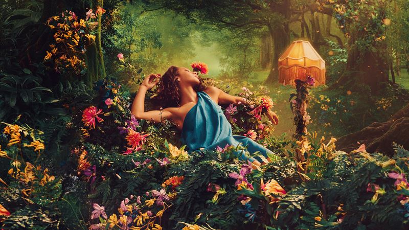 Enjoy a fresh adaptation of Shakespeare's comedy A Midsummer Night's Dream at the Regent's Park Open Air Theatre this summer. Image courtesy of Feast Creative. A young woman wearing a blue, draped dress lies down in a forest surrounded by colorful flowers and plants.