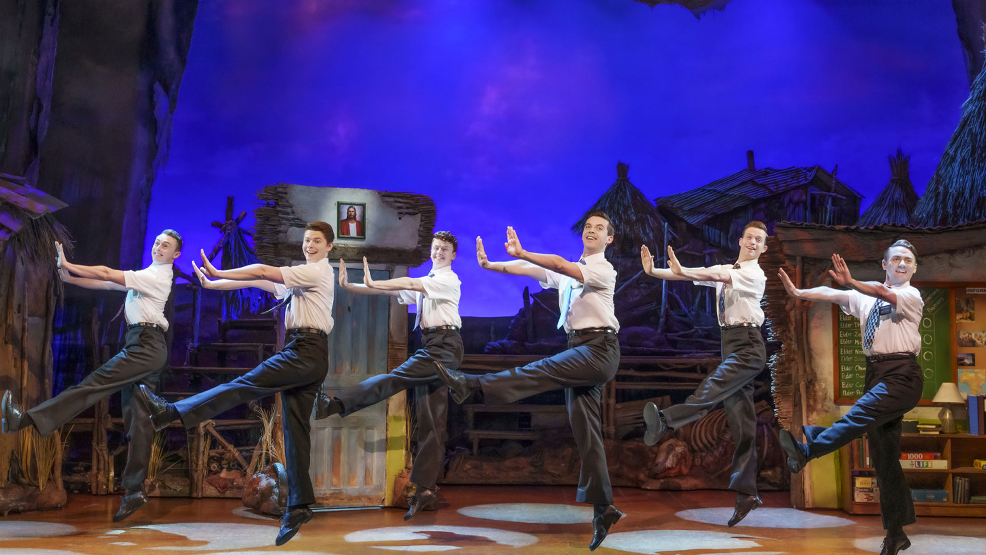 The book of Mormon cast on the London West End Stage at the Prince of Wales Theatre. Image courtesy of London Theatre Direct. The cast of The Book of Mormon is dancing on stage in front of a dark blue background.