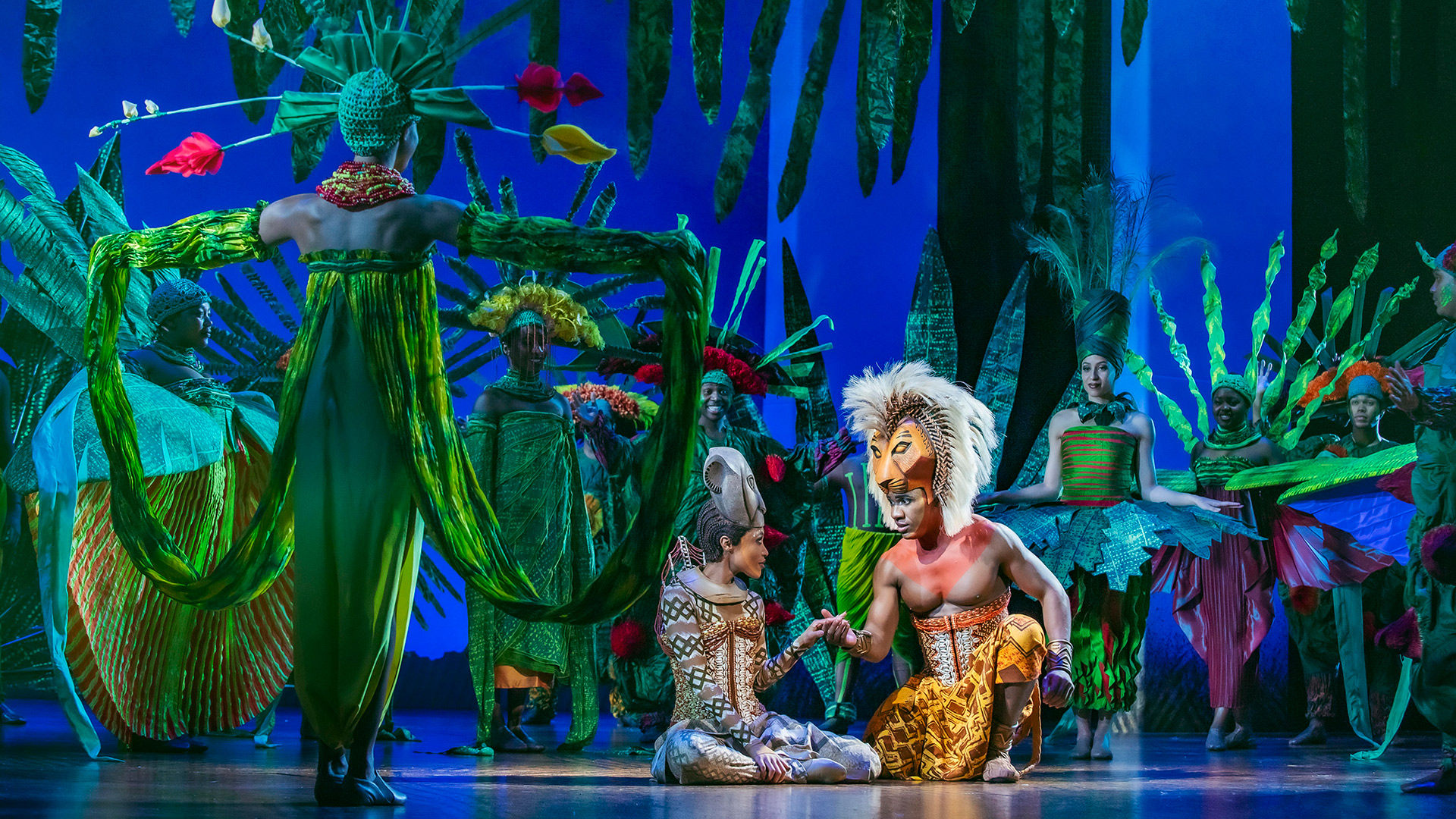 Revisit one of Disney's best-loved animated films, The Lion King, with its long-running musical theatre production, currently at the Lyceum Theatre. Image courtesy of Disney. Actors portraying Simba and Nala hold hands while other members of The Lion King musical cast surround them, dressed and posing as luxurious and tropical trees and plants.