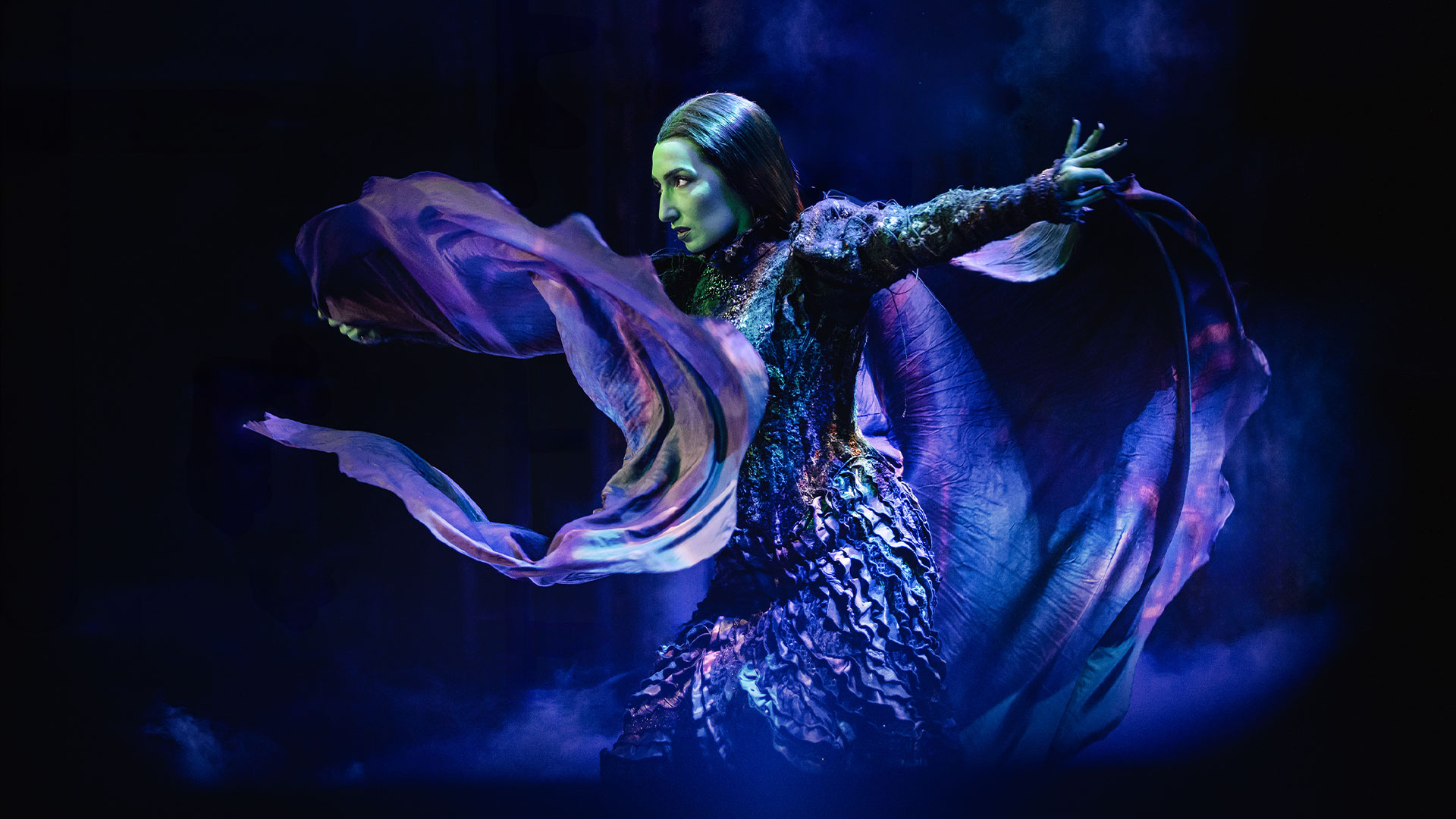 Travel to the world of Oz and discover the story of the unlikely friendship between two witches, Glinda and Elphaba, in Wicked the Musical. Image courtesy of Mark Senior. Elphaba, the Wicked Witch of the West, is shown on stage, opening her arms with her clothes flying around her.