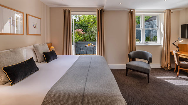 Spacious room at The Resident Kensington. Credit: The Resident Kensington. Image courtesy of The Resident Kensington. A large bed in the foreground with dark grey cushions and a light grey throw, an armchair and desk on the right and open windows with greenery in the background.
